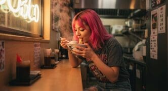 A woman with pink hair smiles as she eats ramen in a restaurant.