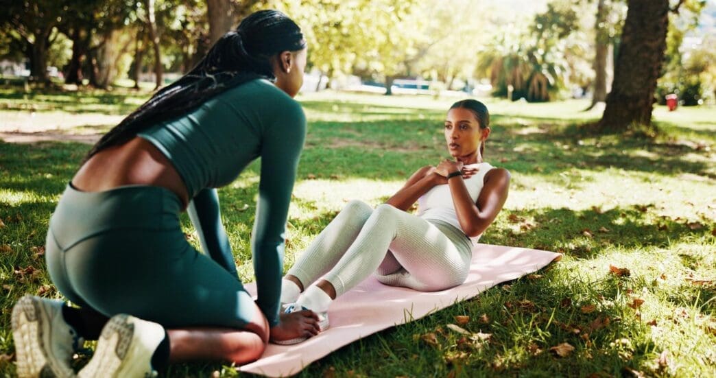 Two women exercise on mats in a park, one coaching the other with a focus on core strength and fitness.