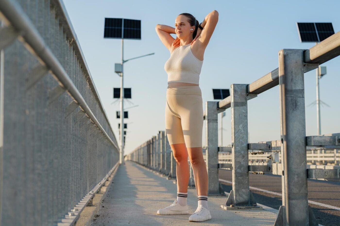A young woman in athletic wear stretches on a sunny bridge, with solar panels visible in the background.