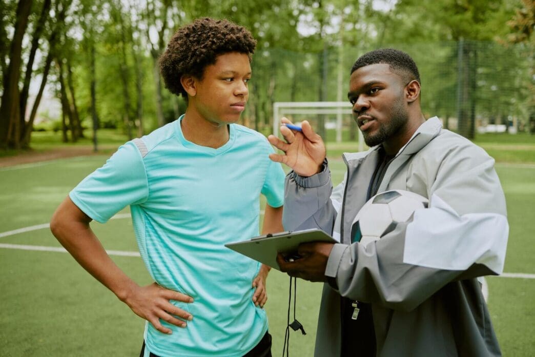 A young boy attentively listens to a black coach explaining soccer strategy on an outdoor field.