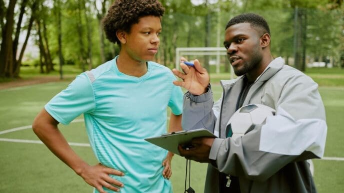 A young boy attentively listens to a black coach explaining soccer strategy on an outdoor field.