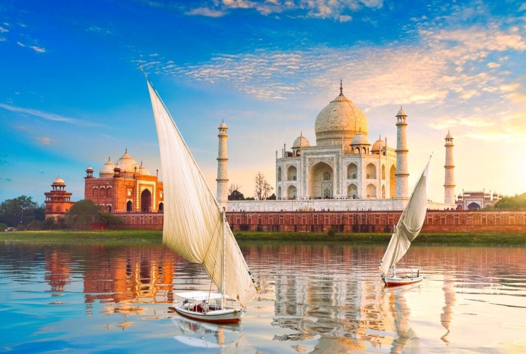 View of the Taj Mahal complex at sunset from the Yamuna River, featuring two sailboats in the foreground and golden light.