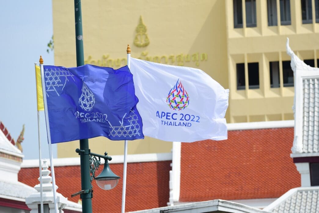Two APEC 2022 Thailand summit flags, one blue and one white, wave on a flagpole in front of buildings in Bangkok.