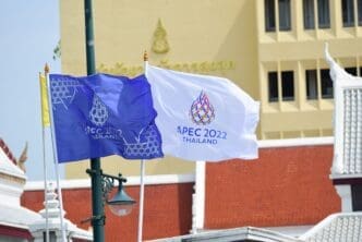 Two APEC 2022 Thailand summit flags, one blue and one white, wave on a flagpole in front of buildings in Bangkok.