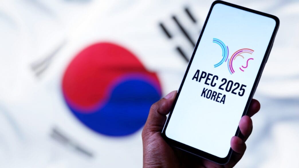 A smartphone displays the APEC 2025 Korea logo, with the South Korean flag blurred behind.