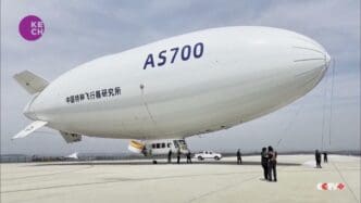Large white airship labeled "AS700" on an airfield with people and a vehicle