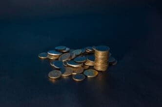 A stack of gold coins sits next to a loose pile of coins on a dark surface.