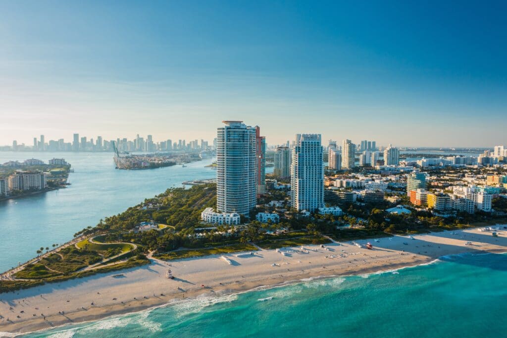 An aerial drone shot of Miami's South Beach, with its sandy shore, high-rise buildings, and the downtown skyline in the distance.