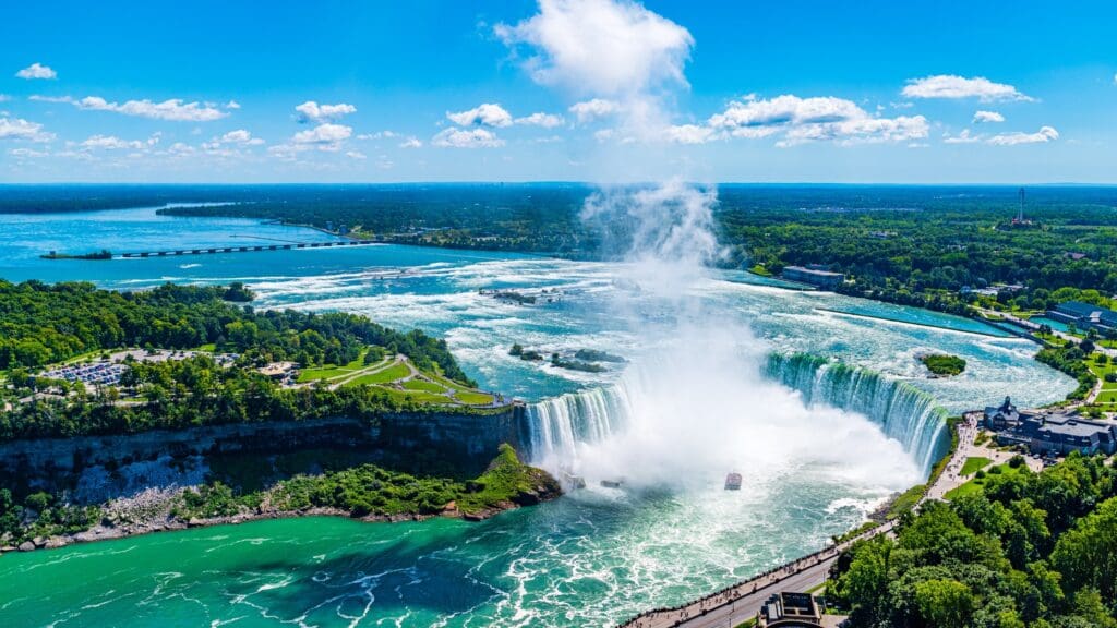 An aerial view of Niagara Falls on a sunny day, showing the wide Horseshoe Falls and a tour boat in the mist below.