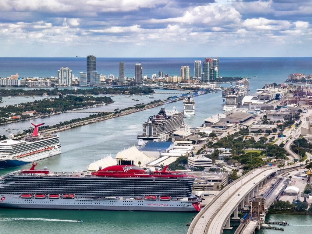 An aerial view shows several cruise ships, including a prominent red Virgin Voyages liner, docked at the Port of Miami, Florida.
