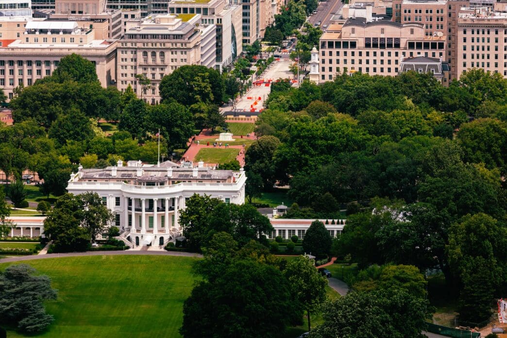 An aerial view shows the White House and its expansive grounds, with the East Wing visible, surrounded by Washington, DC.