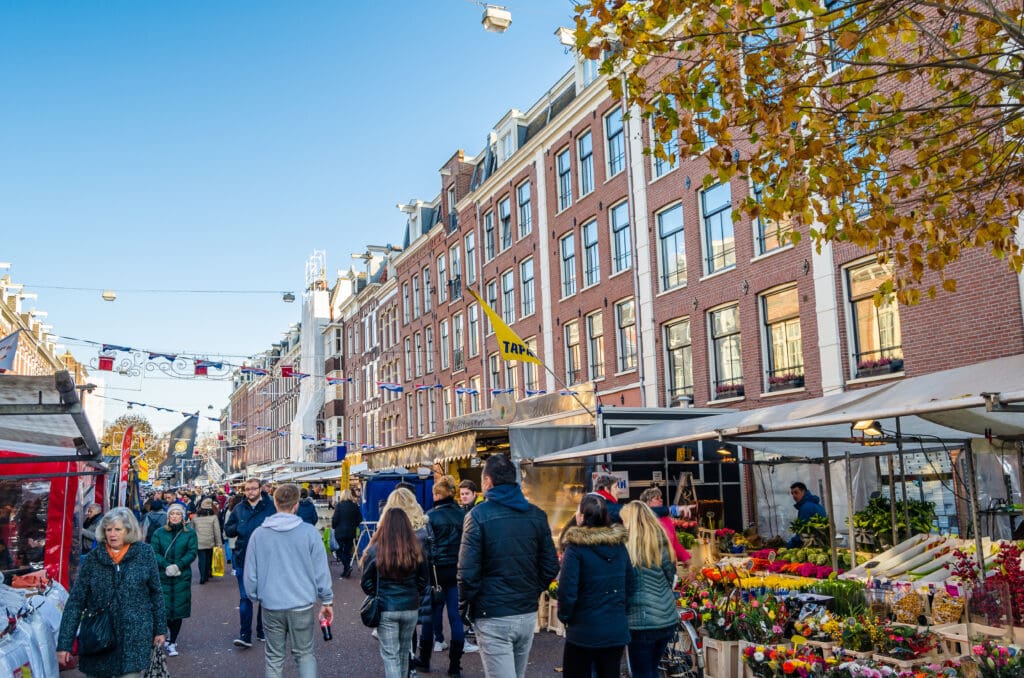 A crowd of shoppers walks through the bustling Albert Cuyp Market in Amsterdam, browsing stalls filled with flowers and produce.