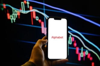 A hand holds a smartphone showing the Alphabet logo in front of a falling stock chart.