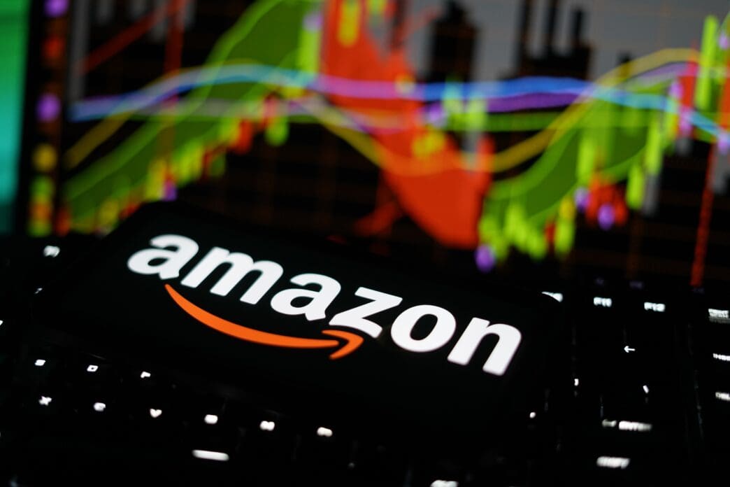 The Amazon logo is displayed on a smartphone, with a colorful, out-of-focus stock market chart in the background.