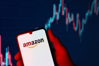 A hand holds a smartphone displaying the Amazon logo against a blurred, red and blue stock chart background.