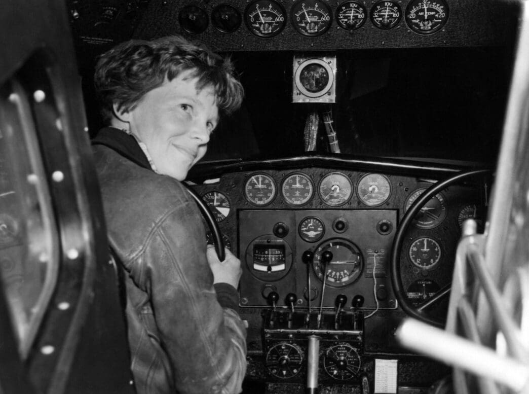 Black and white photo of Amelia Earhart smiling while seated at the complex controls and dashboard of her airplane.