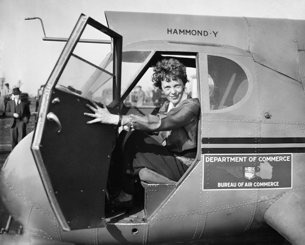 Black and white photo of Amelia Earhart sitting in the cockpit of a "Hammond Y" airplane, smiling with the door open.