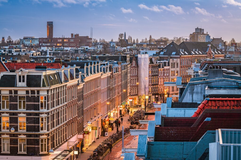A twilight cityscape view of Amsterdam's De Pijp neighborhood shows illuminated streets and rows of historic Dutch houses.