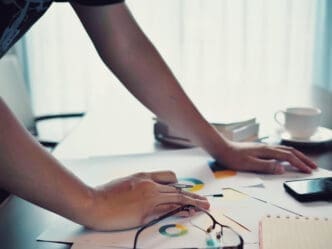 Person standing over a table with printed business charts and scattered papers, analyzing data to create a startup.