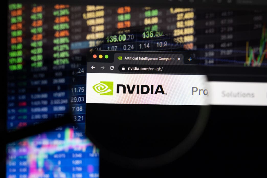 A magnifying glass focuses on the Nvidia logo on its website, with a colorful, out-of-focus stock market chart in the background.