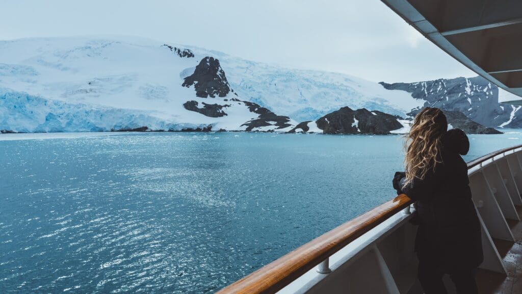 A woman with long hair blowing in the wind stands on a ship's deck railing looking out at an Antarctic glacier and icy blue water.