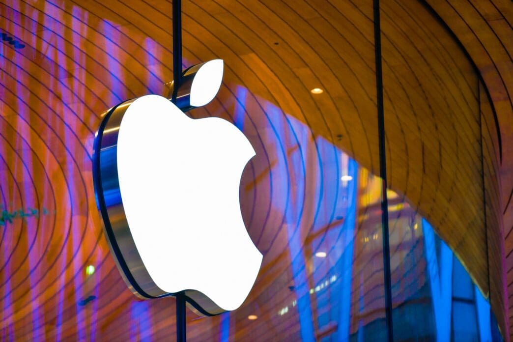 The large, illuminated white Apple logo hangs on the glass facade of the Apple Store Central World in Bangkok, reflecting warm wood and blue neon light.