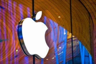 The large, illuminated white Apple logo hangs on the glass facade of the Apple Store Central World in Bangkok, reflecting warm wood and blue neon light.