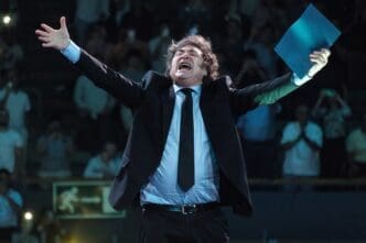 Argentine President Javier Milei with his arms raised in triumph and holding a blue folder, shouting during a conference.