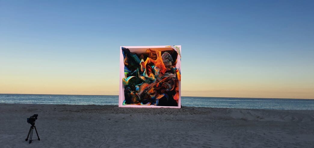 A glowing, colorful, abstract cube sculpture is placed on a sandy beach at sunset, near a tripod-mounted camera.