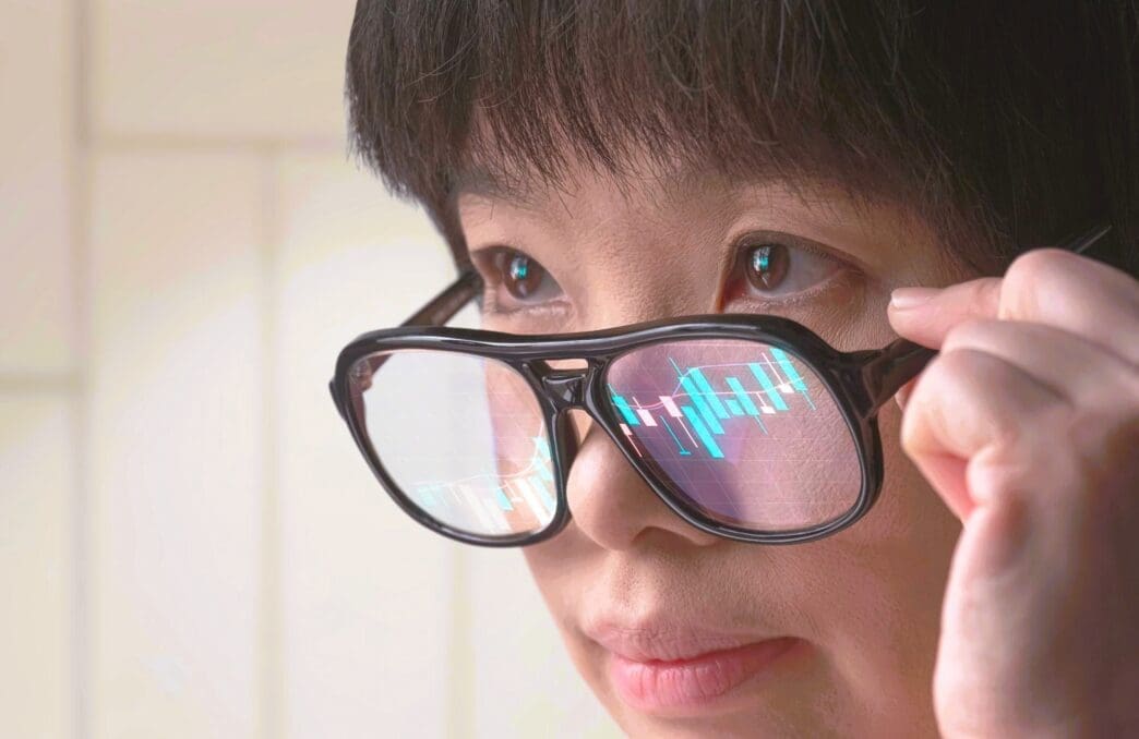 Asian female crypto trader's glasses reflect a blue candlestick stock chart.