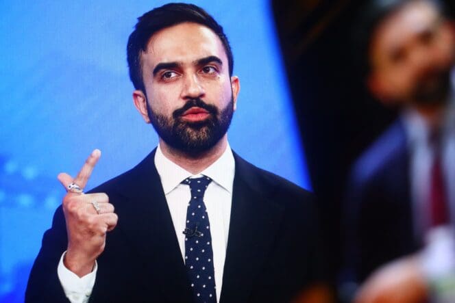 New York State Assemblymember Zohran Kwame Mamdani gestures with one finger while speaking at an event against a bright blue background.