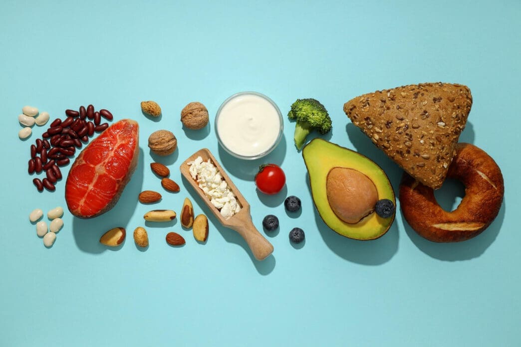 Flat lay of various high-protein, healthy fats, and whole grain foods, including salmon, beans, nuts, yogurt, and avocado, arranged on a blue background.