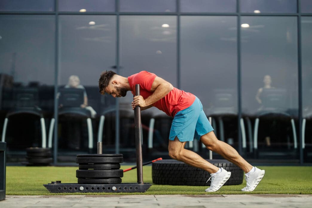 Man in red shirt and blue shorts pushes a heavy black sled on pavement outside a modern gym.