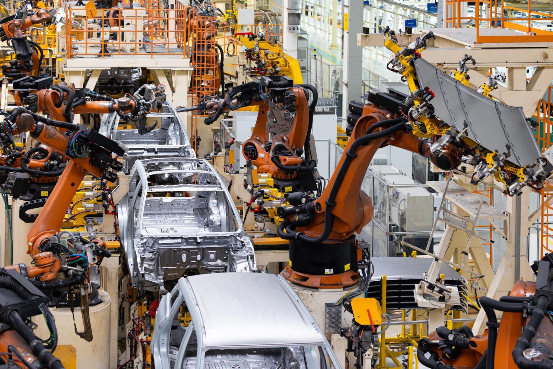 Busy factory floor filled with numerous orange robotic arms working on the assembly of unpainted electric vehicle (EV) body shells.
