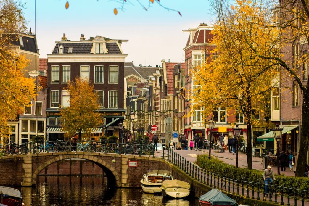 A picturesque Amsterdam canal in autumn, with a brick arch bridge, boats, and historic houses framed by golden leaves.