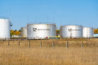 Several large Bashneft oil storage tanks sit behind a fence in a field in Bashkortostan, Russia, under a clear blue sky.