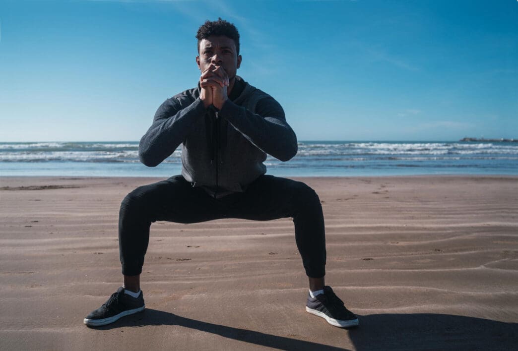 A man in athletic wear performs a squat on a sandy beach with the ocean and sky in the background
