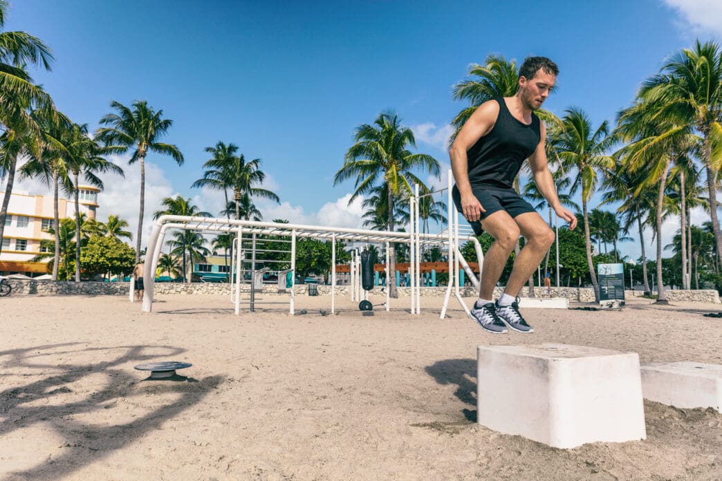 Man in black tank top and shorts jumps over a concrete block on a sandy beach
