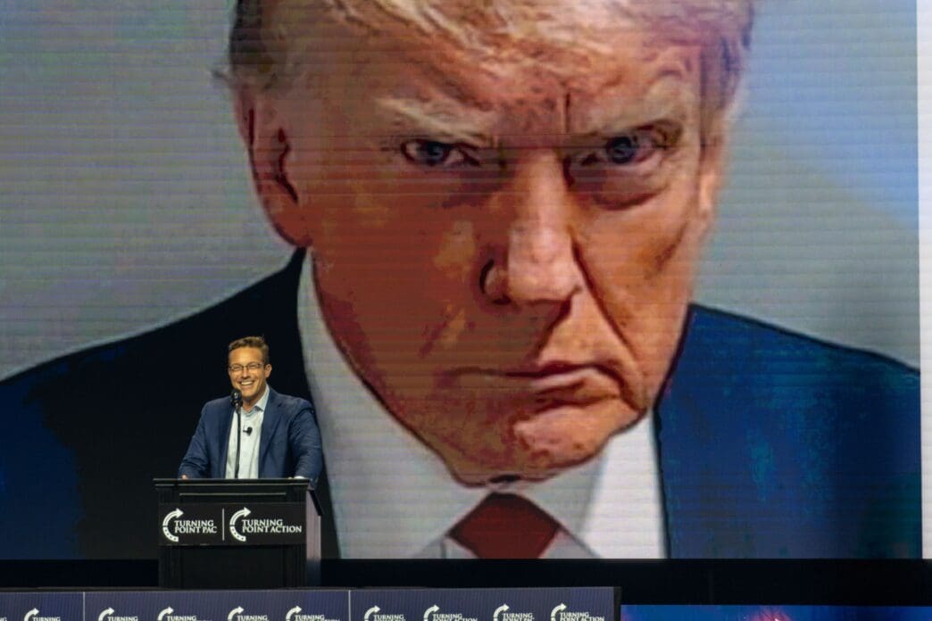 Political commentator Benny Johnson speaking at a podium in front of a massive, close-up screen projection of Donald Trump's face.