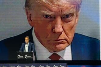 Political commentator Benny Johnson speaking at a podium in front of a massive, close-up screen projection of Donald Trump's face.