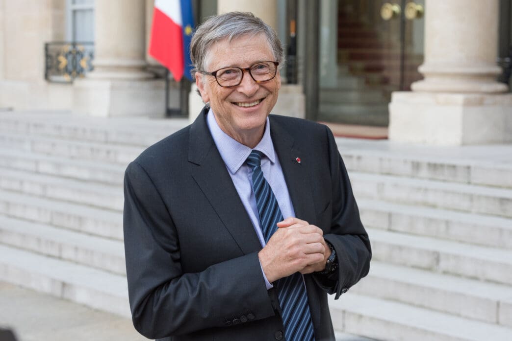 Bill Gates smiling in a dark suit and glasses, standing on the steps outside the official French presidential palace.