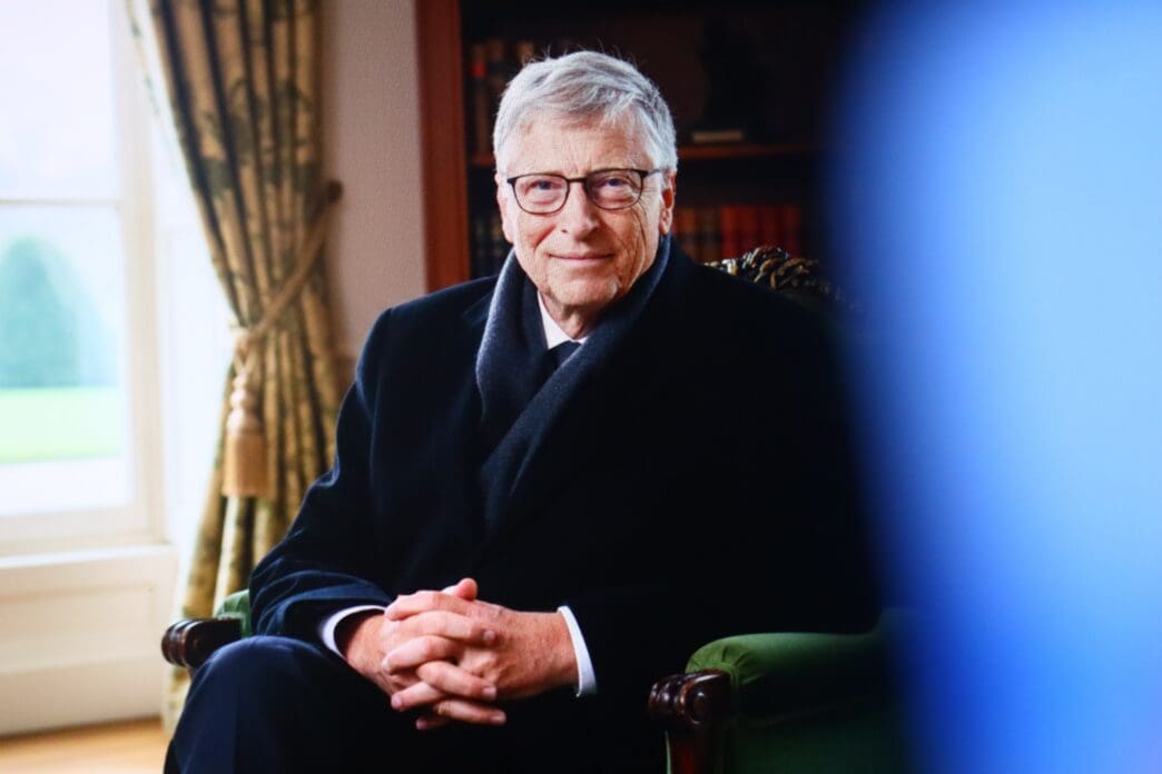Bill Gates, wearing a dark overcoat and glasses, is seated in a green armchair in a formal room, smiling at the camera.