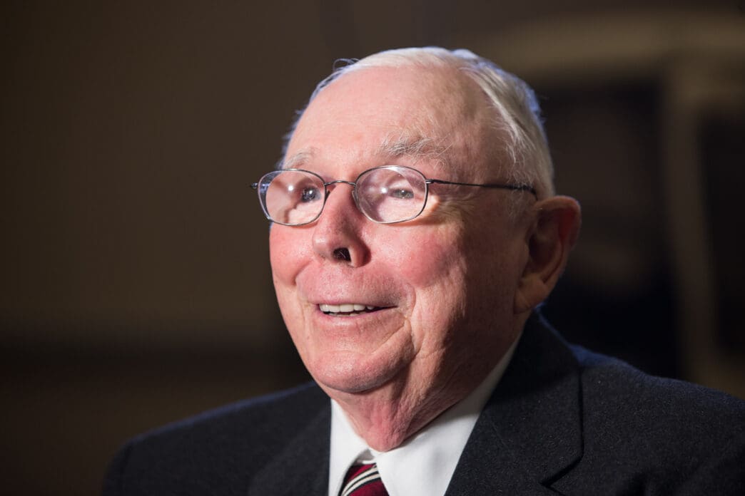 Close-up portrait of Charlie Munger, an elderly man in a suit and glasses, smiling warmly during an interview
