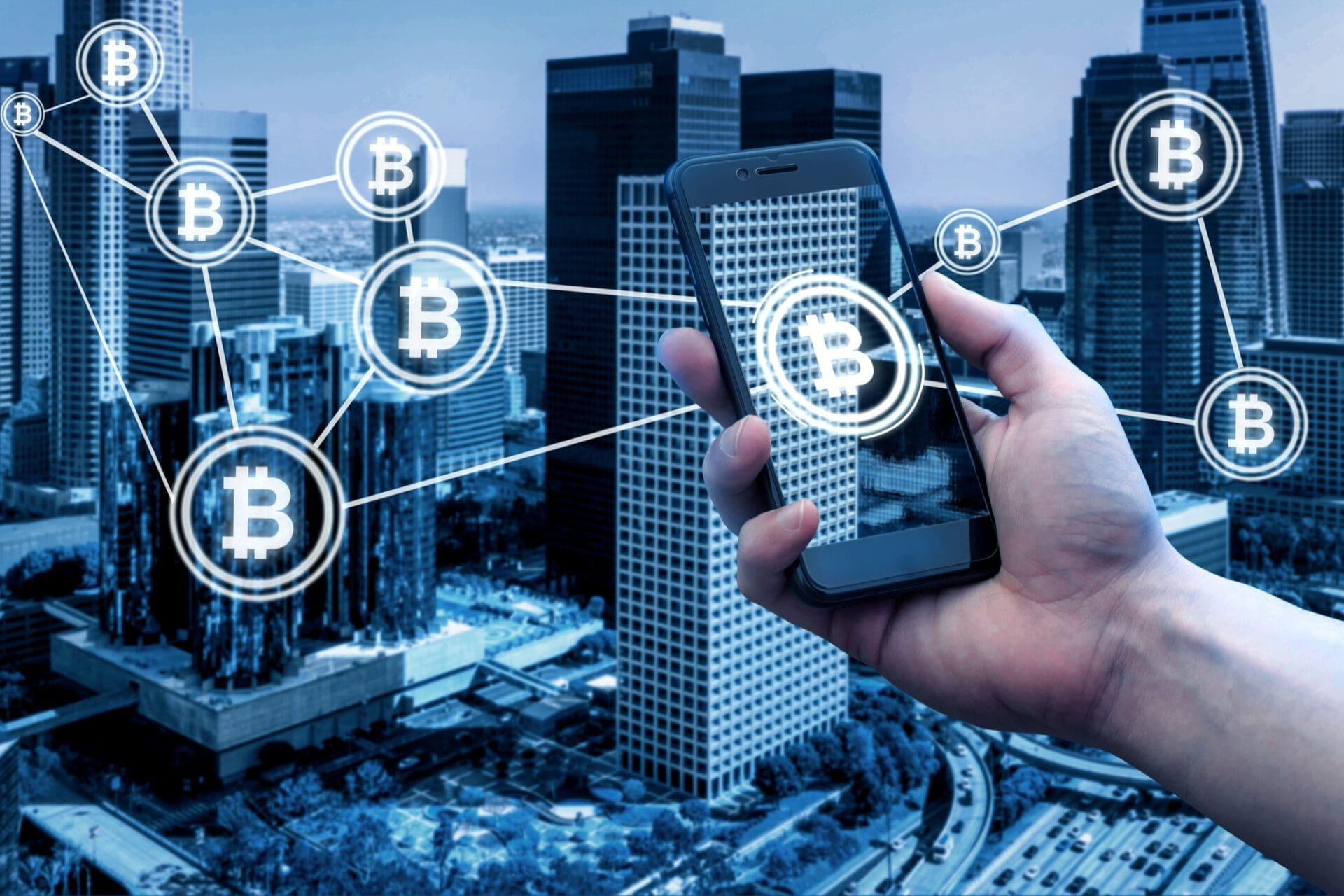 A hand holds a smartphone displaying the Bitcoin logo, overlaid on a cityscape with Bitcoin icons connected by lines