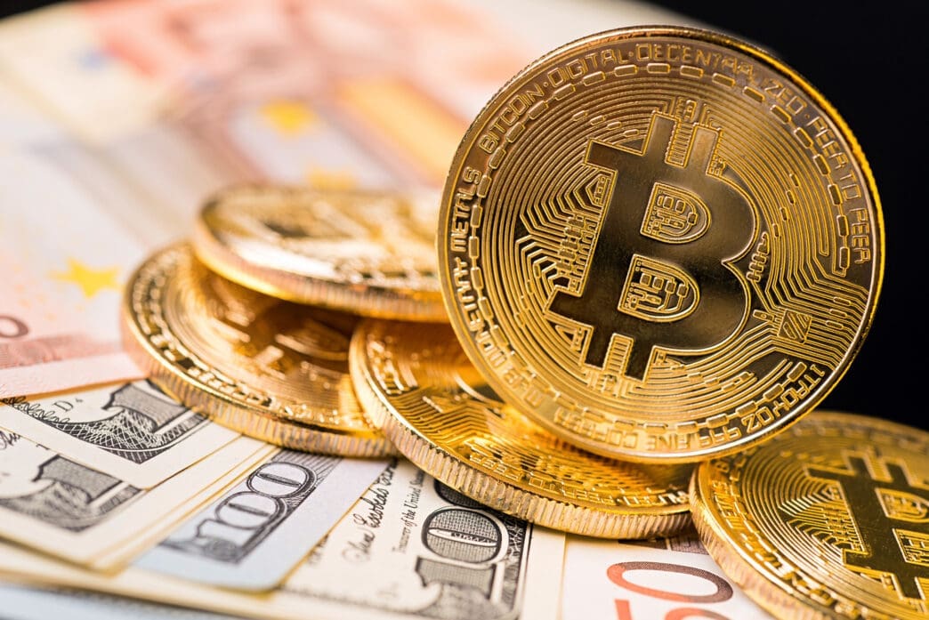 A close-up shot of several gold Bitcoin coins resting on a bed of US dollar and Euro banknotes.