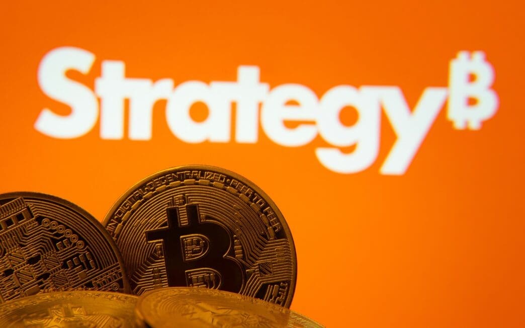 A pile of physical Bitcoin coins with the MicroStrategy logo displayed on an orange screen in the background.