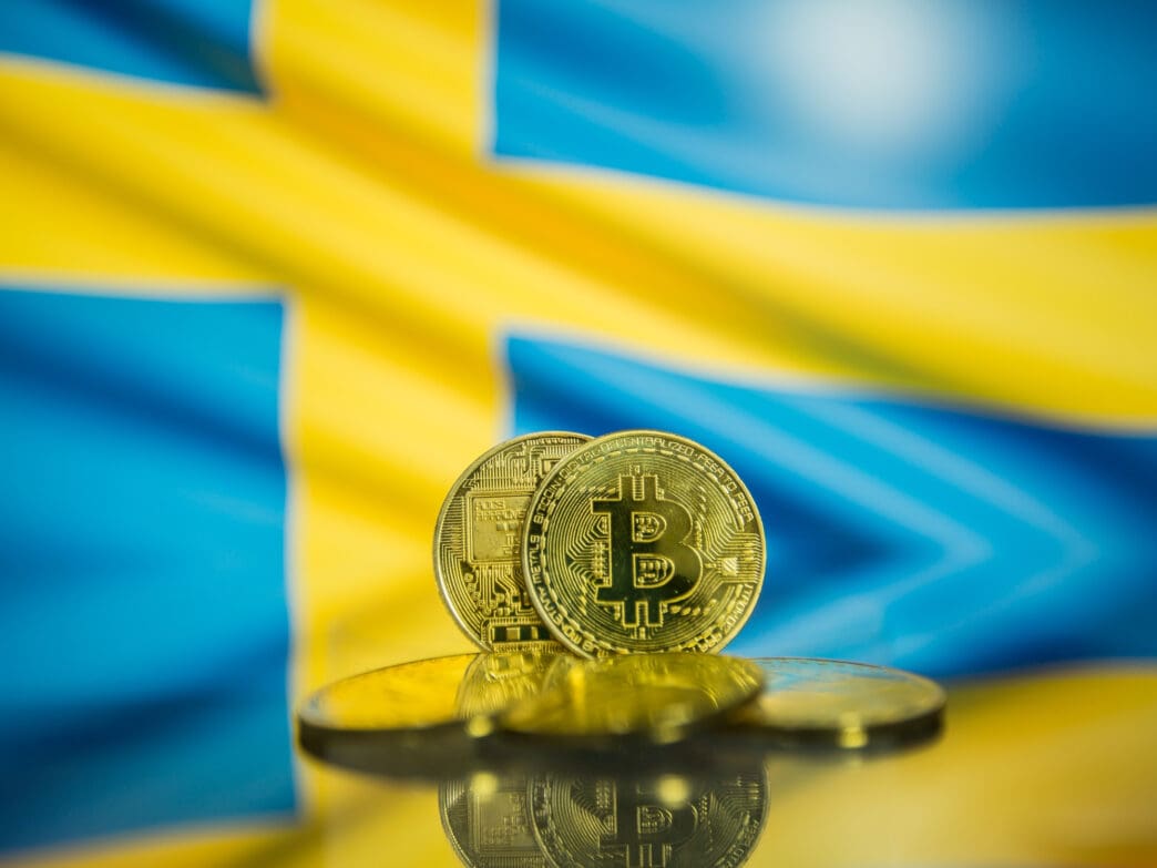 Gold Bitcoin coins rest on a reflective surface with the Swedish flag blurred in the background