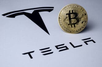 A physical gold Bitcoin coin rests on a white document showing the black Tesla wordmark and logo.