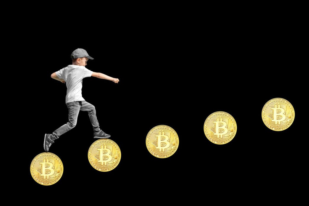 A boy in a cap and casual clothes leaps over a series of Bitcoin coins against a black background
