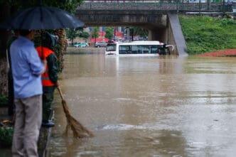 A white bus is half-submerged in a severely flooded urban underpass while people stand under an umbrella watching the scene.